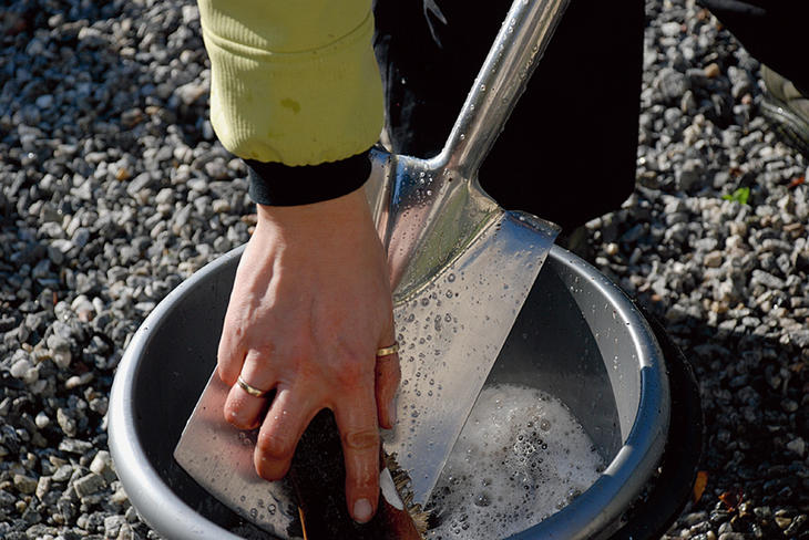 En dame vasker en spade med kost i en grå bøtte. 