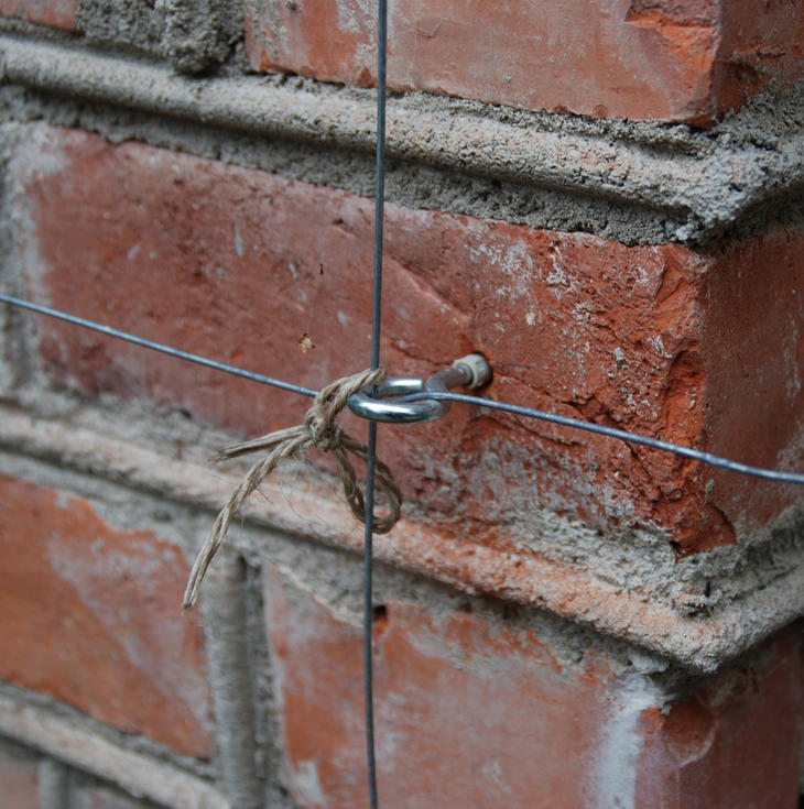 En øyeskrue montert på en murvegg. Gjennom skruen går to ståltråder i kryss. 