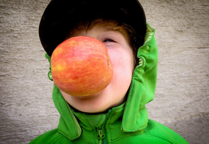 En gutt med grønn jakke holder et stort, rødt eple med munnen. 